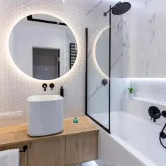 Moderno bagno cieco idee: lavabo bianco su mobile in legno, specchio rotondo illuminato, vasca da bagno con doccia e finiture nere.