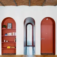 Nicchia nel muro con mensole color terracotta, piena di libri e oggetti decorativi. Tre archi bianchi incorniciano la nicchia, un corridoio e una porta rossa.