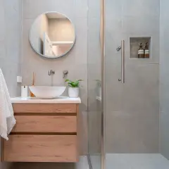 Bagno piccolo con lavabo a conca, mobile in legno, specchio rotondo e doccia con piastrelle grigie.