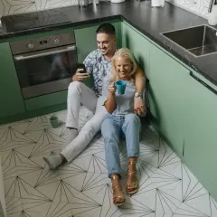 Coppia rilassata in cucina, con mobili verde menta e un pavimento geometrico. Lui guarda il telefono, lei sorseggia una bevanda.