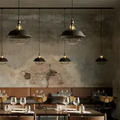 Un elegante lampadario a binario con sei luci industriali illumina un ristorante con pareti in cemento grezzo e divanetti in pelle marrone.
