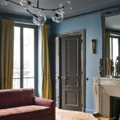 Soggiorno elegante con divano bordeaux, pareti blu balena e tendaggi dorati. Un camino in marmo e sculture africane completano l'arredamento, creando abbinamenti sofisticati.