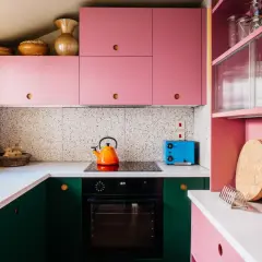 Cucina rosa cipria con mobili verdi, piano cottura, tostapane blu e decorazioni in terracotta.
