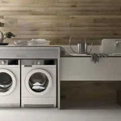 Lavatrice in cucina moderna, con due elettrodomestici bianchi affiancati sotto un piano di lavoro con bonsai e asciugamani.