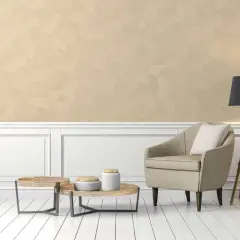 Poltrona beige e tavolini in legno su pavimento bianco, con parete in color champagne e lampada nera.