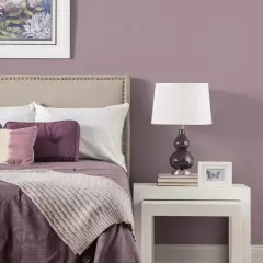Letto con testiera imbottita, cuscini viola e una coperta color malva. Sul comodino, una lampada e una cornice con una farfalla.