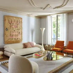 Soggiorno moderno con divano bianco, poltrone arancioni e arazzo colorato appeso alle pareti color crema.