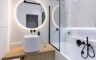 Moderno bagno cieco idee: lavabo bianco su mobile in legno, specchio rotondo illuminato, vasca da bagno con doccia e finiture nere.