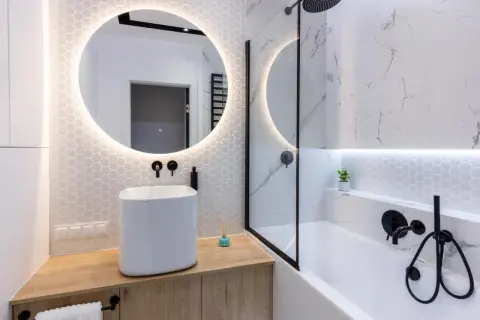 Moderno bagno cieco idee: lavabo bianco su mobile in legno, specchio rotondo illuminato, vasca da bagno con doccia e finiture nere.