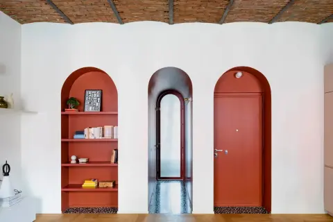 Nicchia nel muro con mensole color terracotta, piena di libri e oggetti decorativi. Tre archi bianchi incorniciano la nicchia, un corridoio e una porta rossa.
