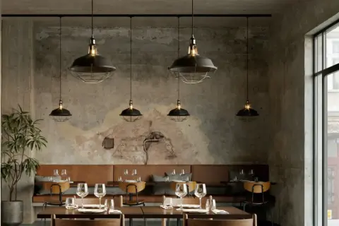 Un elegante lampadario a binario con sei luci industriali illumina un ristorante con pareti in cemento grezzo e divanetti in pelle marrone.