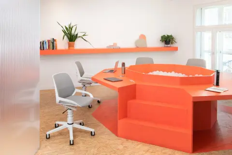 Un ufficio moderno con un tavolo arancione a forma di esagono, dotato di una vasca di palline e sedie ergonomiche.