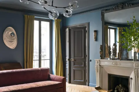 Soggiorno elegante con divano bordeaux, pareti blu balena e tendaggi dorati. Un camino in marmo e sculture africane completano l'arredamento, creando abbinamenti sofisticati.