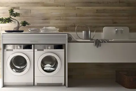 Lavatrice in cucina moderna, con due elettrodomestici bianchi affiancati sotto un piano di lavoro con bonsai e asciugamani.