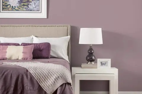 Letto con testiera imbottita, cuscini viola e una coperta color malva. Sul comodino, una lampada e una cornice con una farfalla.