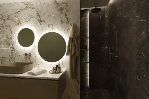 Elegante bagno scuro moderno con lavabo in pietra, specchi rotondi illuminati e doccia in marmo scuro.