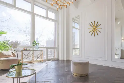 Un elegante soggiorno con un divano beige, tavolini rotondi in vetro e oro, e un orologio da parete dorato. Grigio e oro stanno bene insieme, creando un'atmosfera sofisticata.
