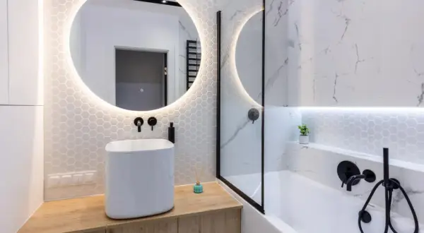 Moderno bagno cieco idee: lavabo bianco su mobile in legno, specchio rotondo illuminato, vasca da bagno con doccia e finiture nere.