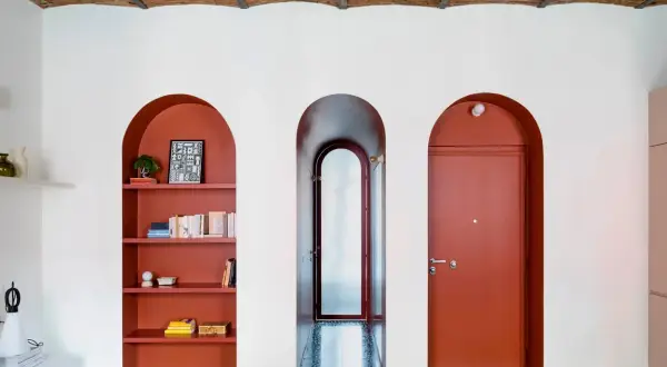 Nicchia nel muro con mensole color terracotta, piena di libri e oggetti decorativi. Tre archi bianchi incorniciano la nicchia, un corridoio e una porta rossa.