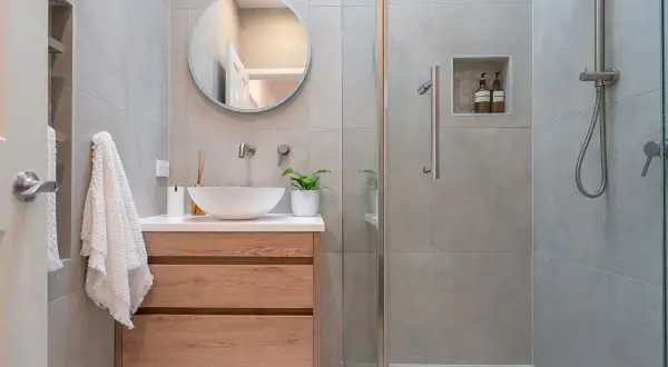 Bagno piccolo con lavabo a conca, mobile in legno, specchio rotondo e doccia con piastrelle grigie.