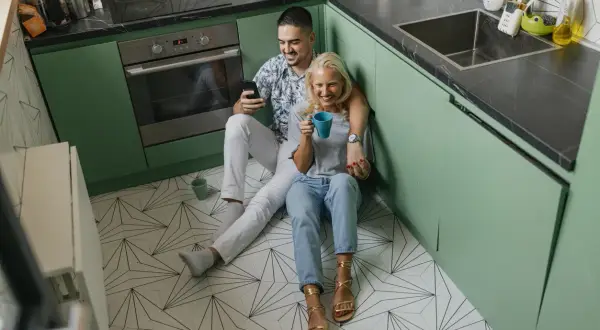 Coppia rilassata in cucina, con mobili verde menta e un pavimento geometrico. Lui guarda il telefono, lei sorseggia una bevanda.