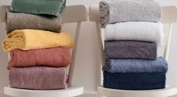 Ecco come piegare gli asciugamani in modo elegante: pile di asciugamani colorati e neutri disposti su sedie bianche.