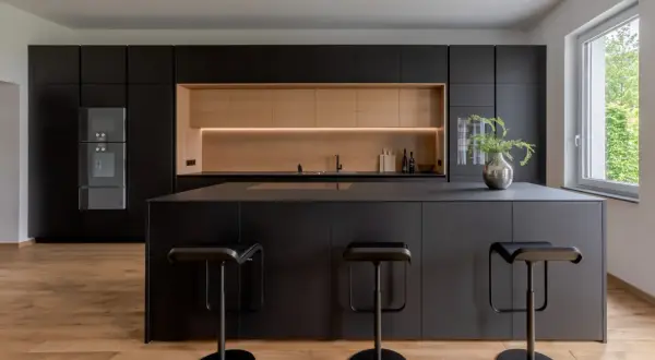 Moderna cucina nera con isola centrale e sgabelli. Un'elegante nicchia in legno chiaro illumina lo spazio, offrendo splendide idee cucina nera e legno.