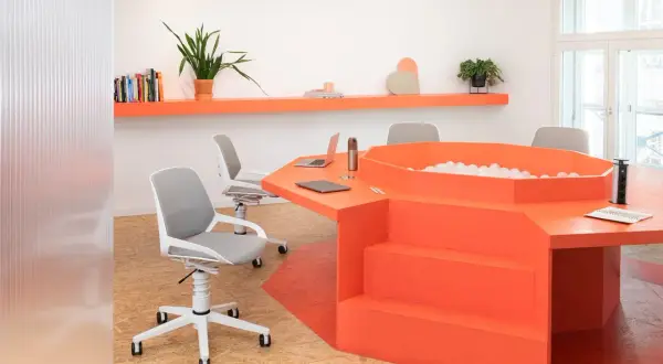 Un ufficio moderno con un tavolo arancione a forma di esagono, dotato di una vasca di palline e sedie ergonomiche.