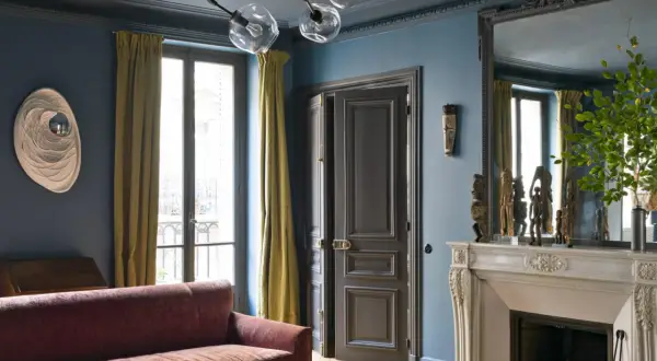 Soggiorno elegante con divano bordeaux, pareti blu balena e tendaggi dorati. Un camino in marmo e sculture africane completano l'arredamento, creando abbinamenti sofisticati.