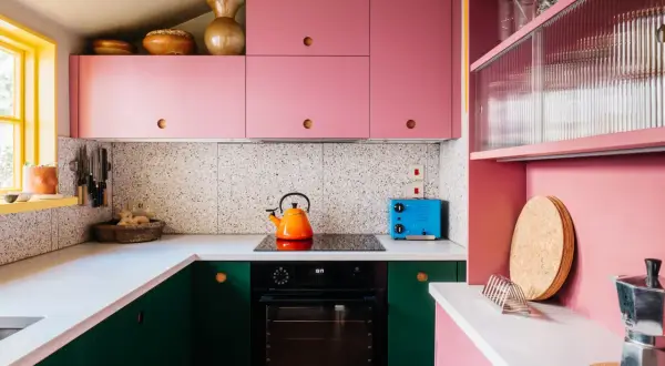 Cucina rosa cipria con mobili verdi, piano cottura, tostapane blu e decorazioni in terracotta.