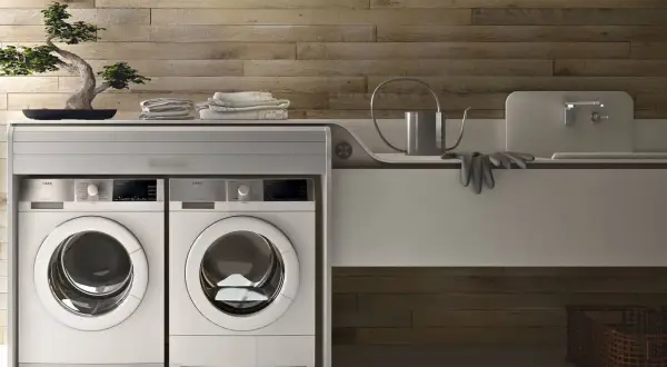 Lavatrice in cucina moderna, con due elettrodomestici bianchi affiancati sotto un piano di lavoro con bonsai e asciugamani.