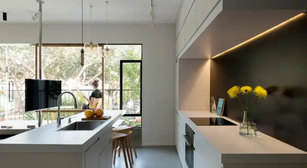 Un moderno cucina soggiorno open space di 50 mq con isola centrale, bancone con lavello e un'area cottura lineare. Una donna legge vicino alla finestra.