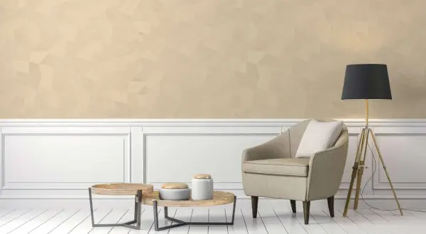 Poltrona beige e tavolini in legno su pavimento bianco, con parete in color champagne e lampada nera.
