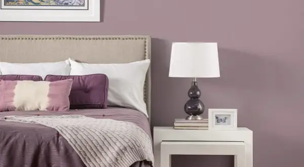 Letto con testiera imbottita, cuscini viola e una coperta color malva. Sul comodino, una lampada e una cornice con una farfalla.