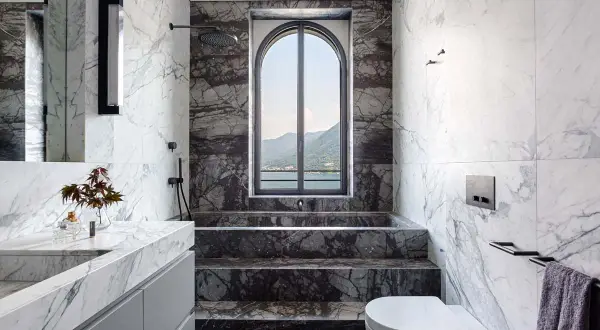 Bagno di lusso con doccia in muratura, vasca e lavabo in marmo, affacciato su un lago e montagne.