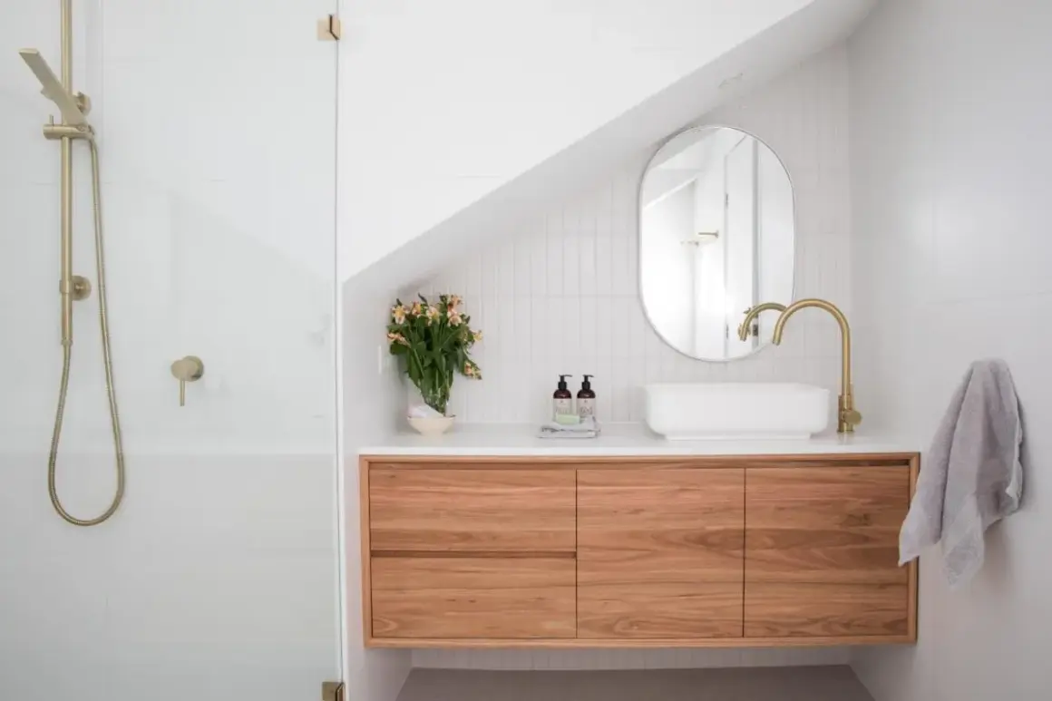 Un bagno moderno con doccia, mobile sospeso in legno, lavabo bianco e rubinetteria oro. Idee per come arredare un bagno con stile.
