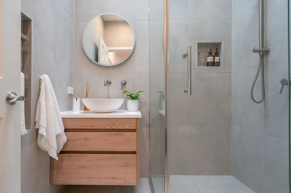 Bagno piccolo con lavabo a conca, mobile in legno, specchio rotondo e doccia con piastrelle grigie.