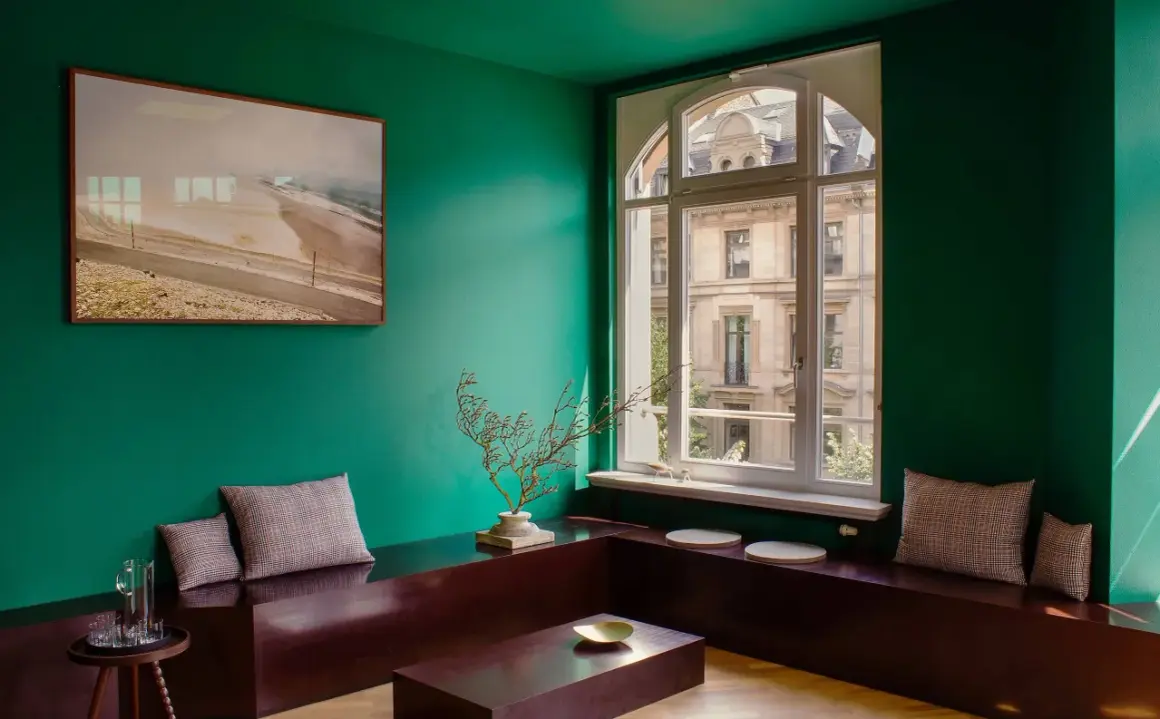 Un soggiorno moderno con pareti verde smeraldo, un quadro astratto, un divano basso in legno scuro e cuscini a quadri. I colori che stanno bene con il verde sono esaltati dal contrasto.