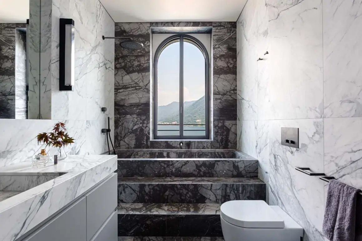 Bagno di lusso con doccia in muratura, vasca e lavabo in marmo, affacciato su un lago e montagne.