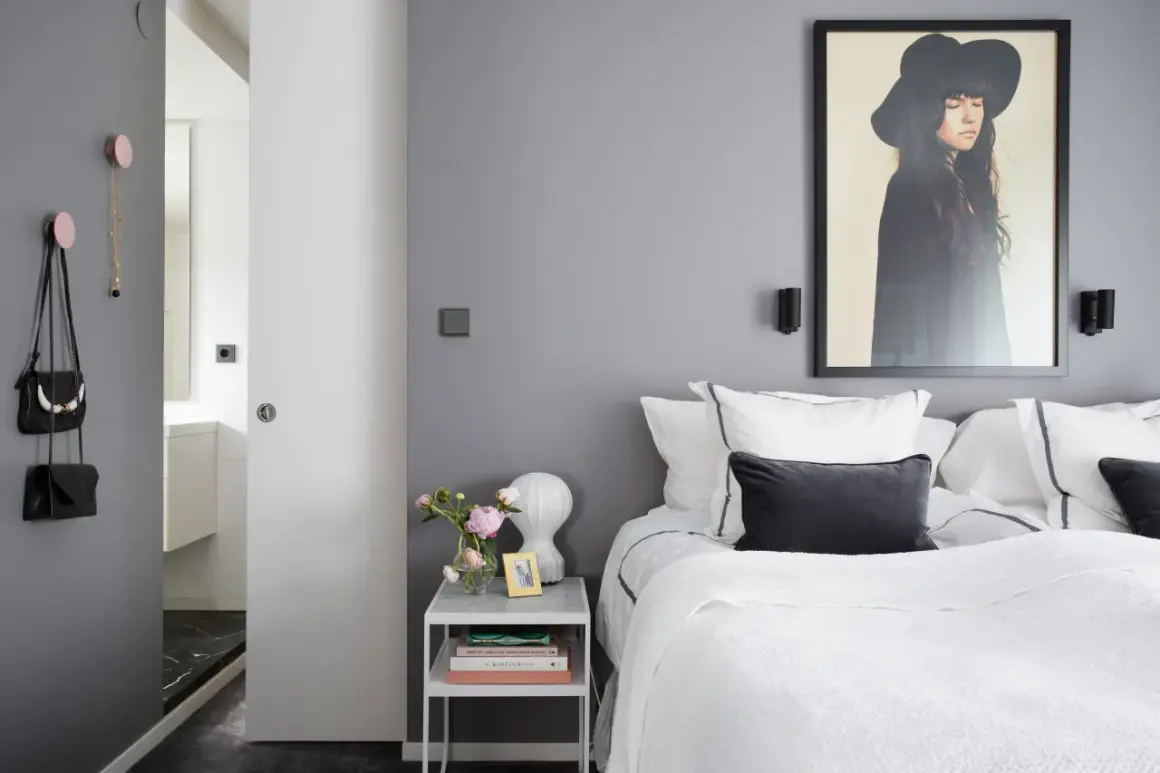 Pareti camera da letto grigia e bianche, con letto bianco, quadro di una donna con cappello e accessori appesi.