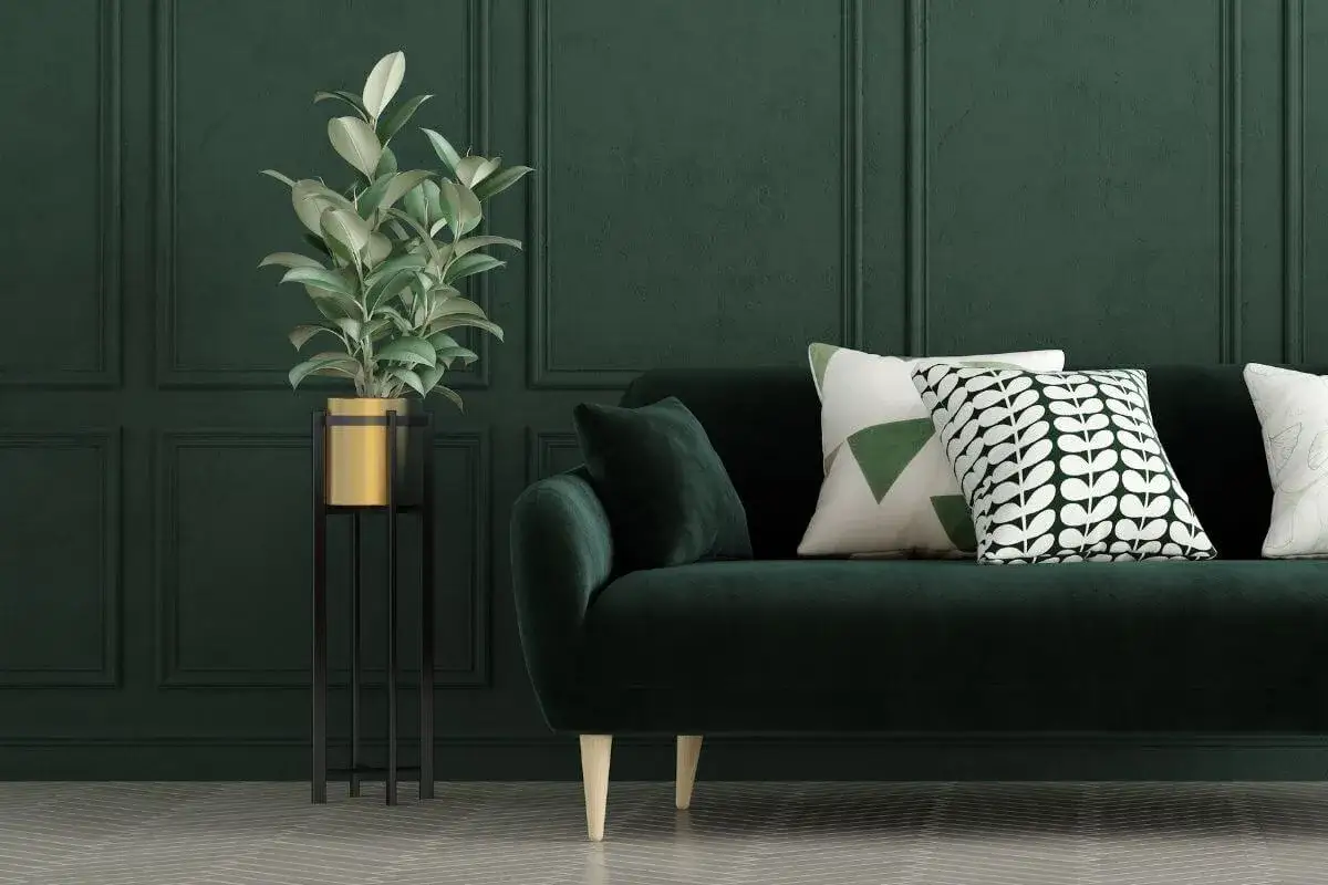 Divano verde smeraldo in una stanza con pareti verde foresta. Una pianta in vaso dorato su un supporto nero. Cuscini decorativi con motivi geometrici e fogliacei.