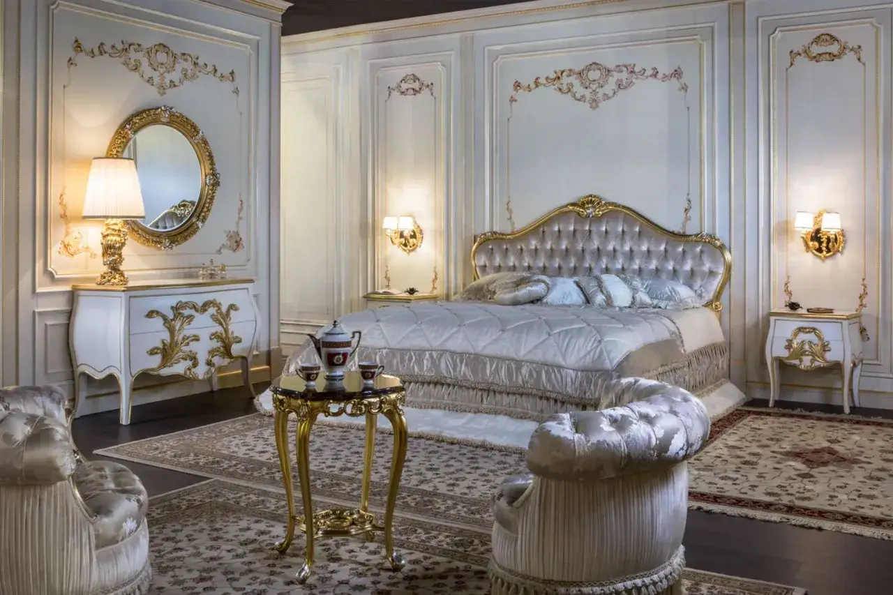 Camera da letto classica: letto capitonné dorato, comò bianco con intagli oro, specchio rotondo, poltrone e tavolino coordinati.