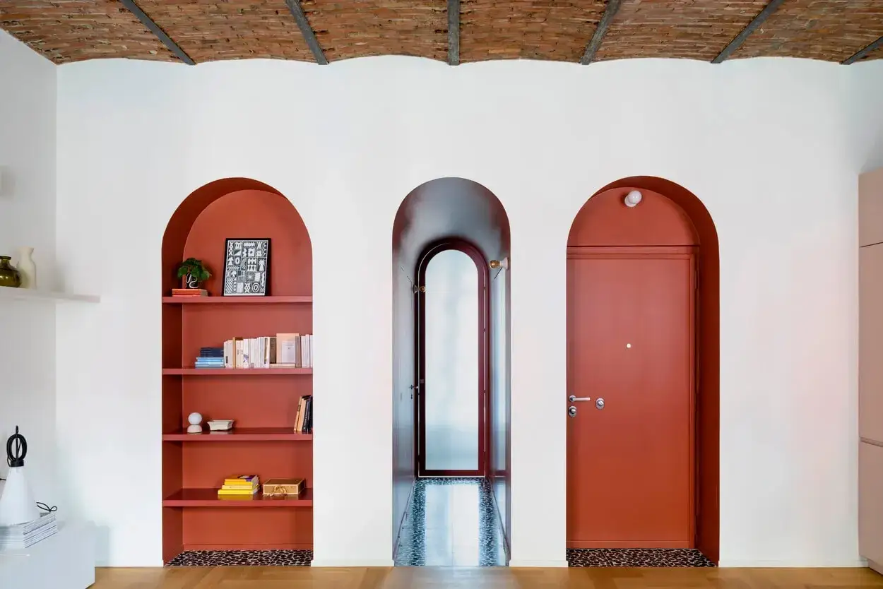 Nicchia nel muro con mensole color terracotta, piena di libri e oggetti decorativi. Un arco rosso conduce a un corridoio con porta e un'altra nicchia con porta rossa.