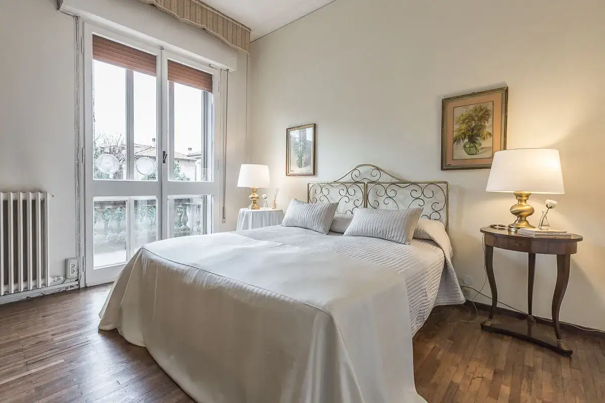 Elegante camera da letto mix antico e moderno con letto bianco, testiera in ferro battuto, comodini in legno e un quadro floreale.