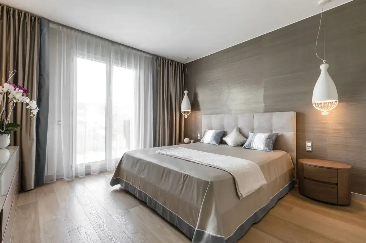 Camera da letto moderna con tendenze di tessuti eleganti. Tende doppie, una pesante color tortora e una leggera in velo bianco, incorniciano una grande finestra.