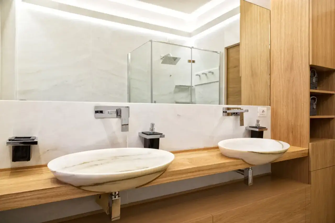 Un bagno elegante e raffinato con doppio lavabo in marmo, rubinetteria cromata e mobili in legno chiaro. La doccia a vista riflette il lusso.