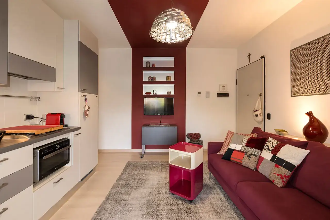Soggiorno moderno con divano e cuscini patchwork, un tavolino bordeaux e bianco, e una cucina a vista. Il colore vinaccia domina il divano e una parete accentuata.