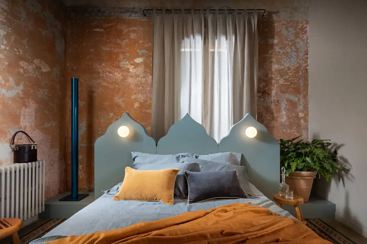 Idee per come rinnovare una vecchia camera da letto: un letto azzurro con testiera a punta, cuscini color ocra e antracite, coperta arancione, pareti arrugginite.