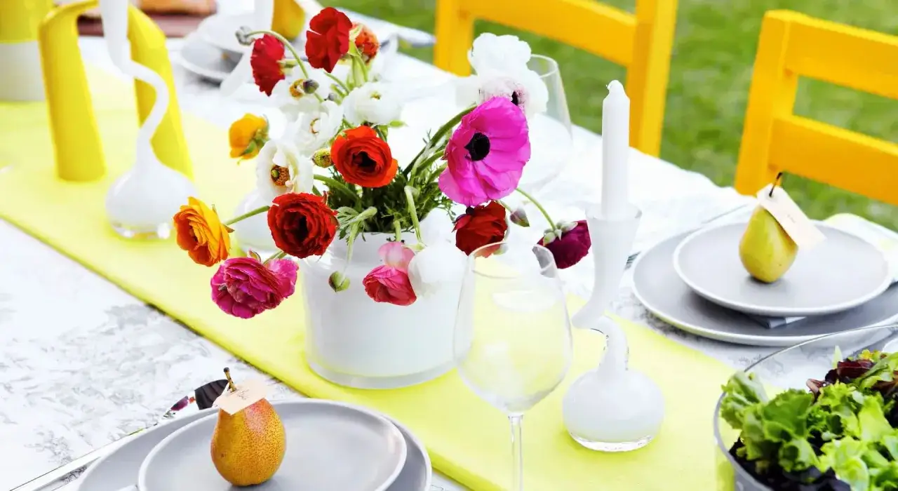 Un tavolo apparecchiato all'aperto con un vaso di fiori colorati, pere come segnaposto e sedie gialle. Idee originali centrotavola fai da te per un'occasione speciale.