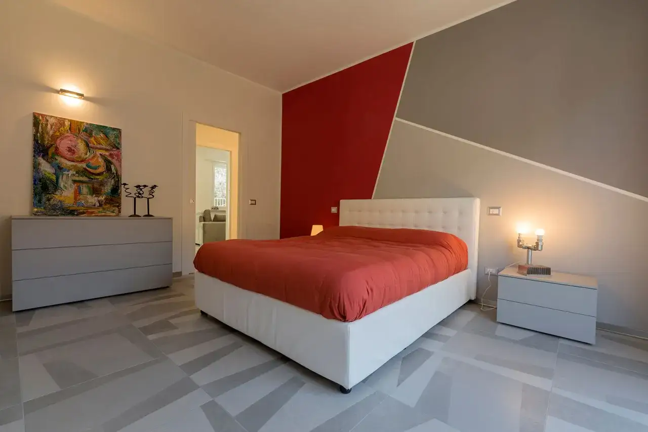 Accogliente zona notte all'ingresso con letto matrimoniale, testiera capitonn&eacute;, copriletto arancione e un quadro astratto.
